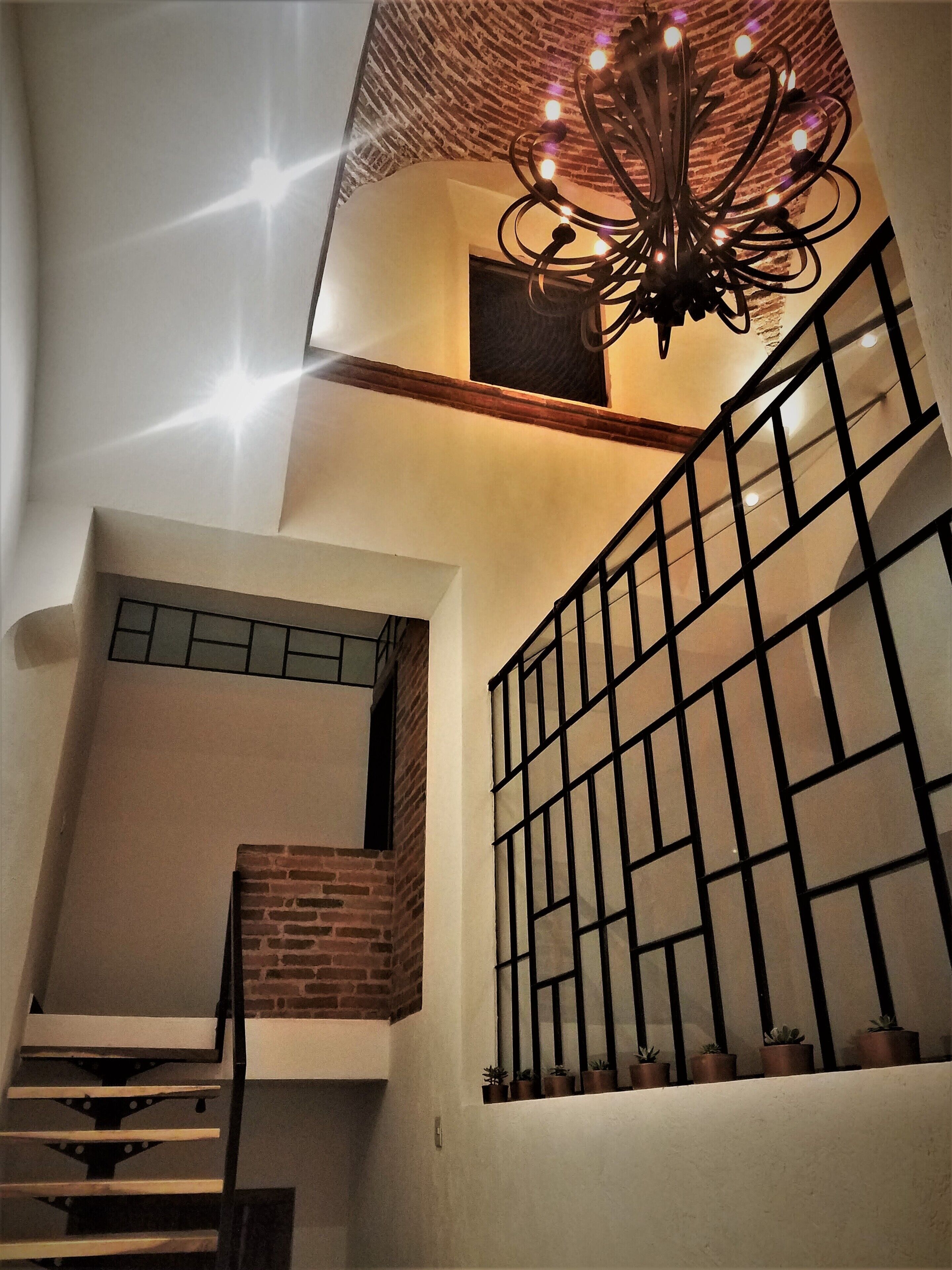 Un escalier avec un mur en briques, une rampe en métal noir et un lustre à plusieurs lampes.