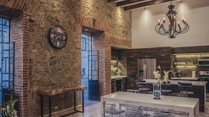 A modern kitchen with a large island, a dining area with a white table and chairs, and a rustic brick wall with a clock. - Luxurious apartment in historic building  downtown Puebla SV311 -12 (PUEBLA)
