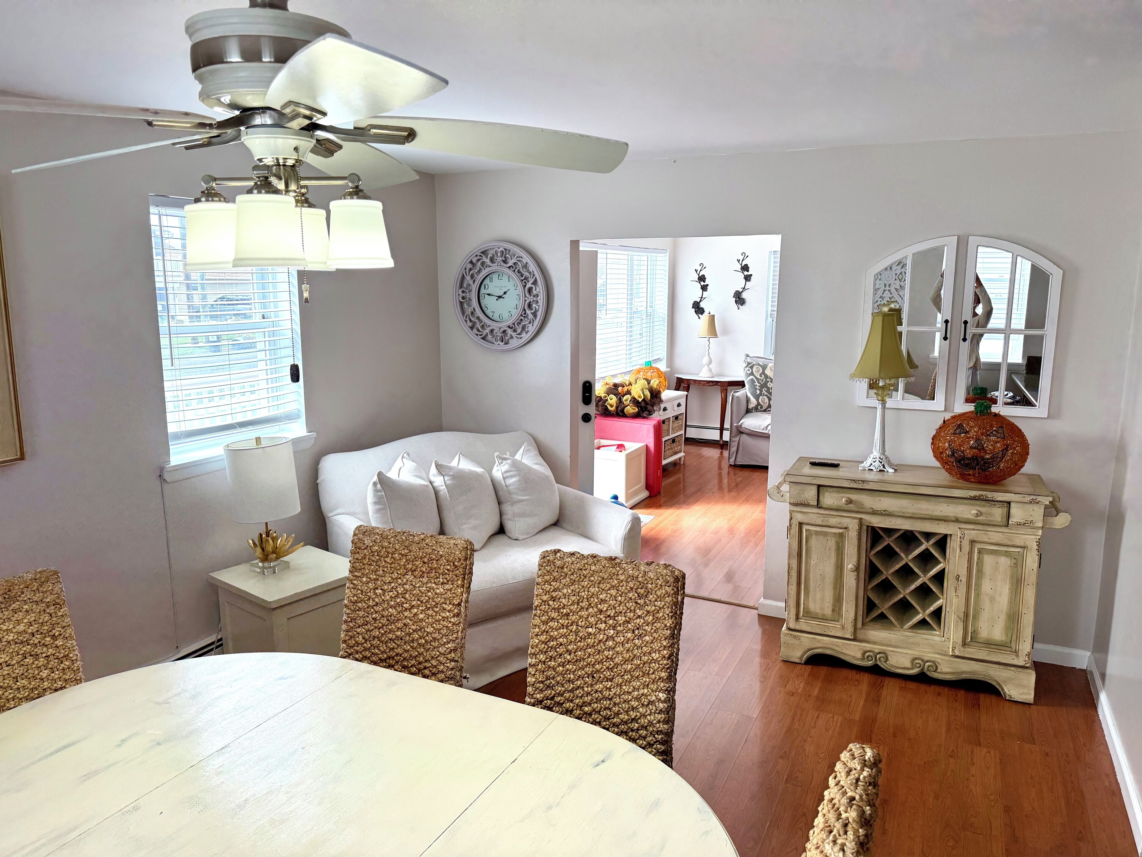 A living room with a white sofa, a dining table, wicker chairs, a ceiling fan, a clock, and a cabinet with a mirror.