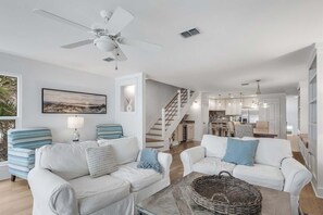 Un amplio salón con un conjunto de sofá blanco, un ventilador de techo y una escalera que lleva a una cocina.