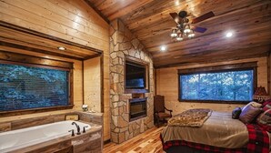 L’intérieur d’une cabane en rondins, avec une cheminée en pierre, une grande fenêtre, un lit recouvert d’une couverture en fourrure et une baignoire.