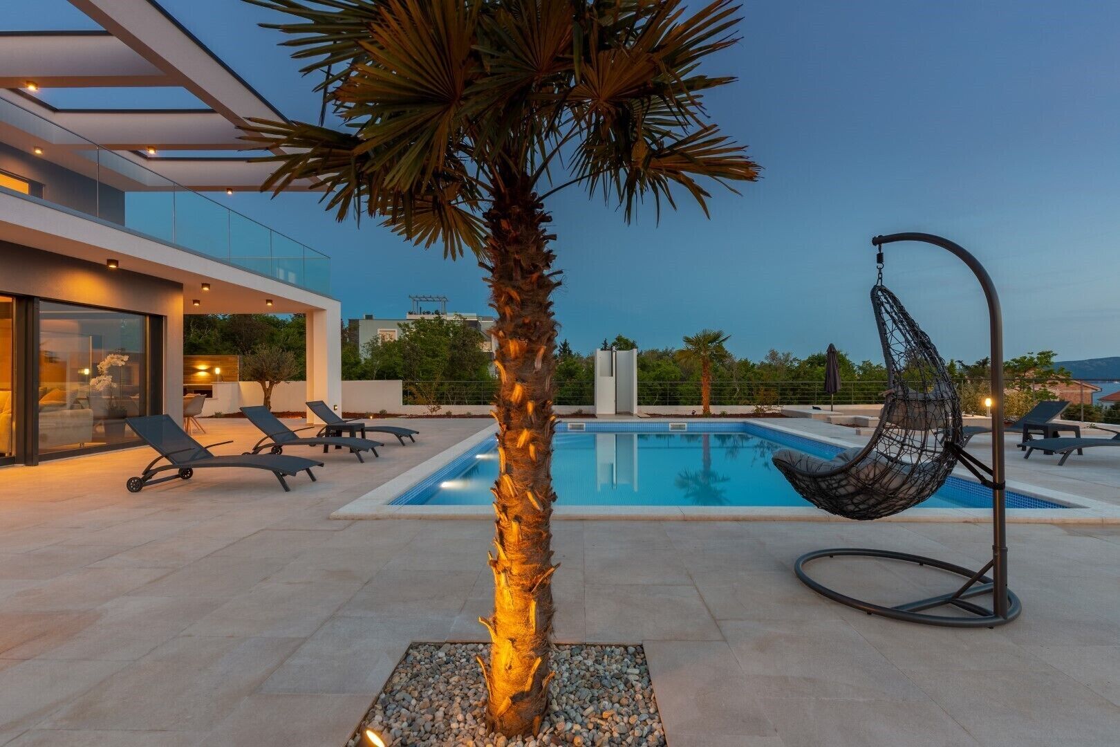 A modern outdoor pool area with a hanging chair, lounge chairs, and a palm tree.