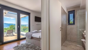 Un dormitorio con cama, un amplio ventanal corredero que da a un balcón, zona de ducha y televisor de pared.