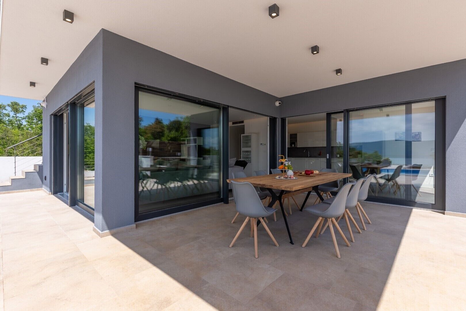A modern outdoor dining area with a wooden table and chairs, large sliding glass doors, and a view of the surrounding landscape.