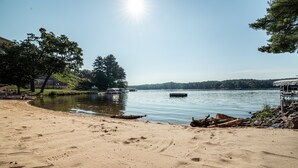 Ein Sandstrand mit Fußabdrücken, ein ruhiger See, ein Boot und ein Steg mit überdachtem Bereich.