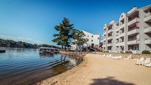 Ein Strand mit einer Reihe weißer Liegen und einer Reihe weißer Gebäude mit Balkonen, ein Baum und eine Anlegestelle.