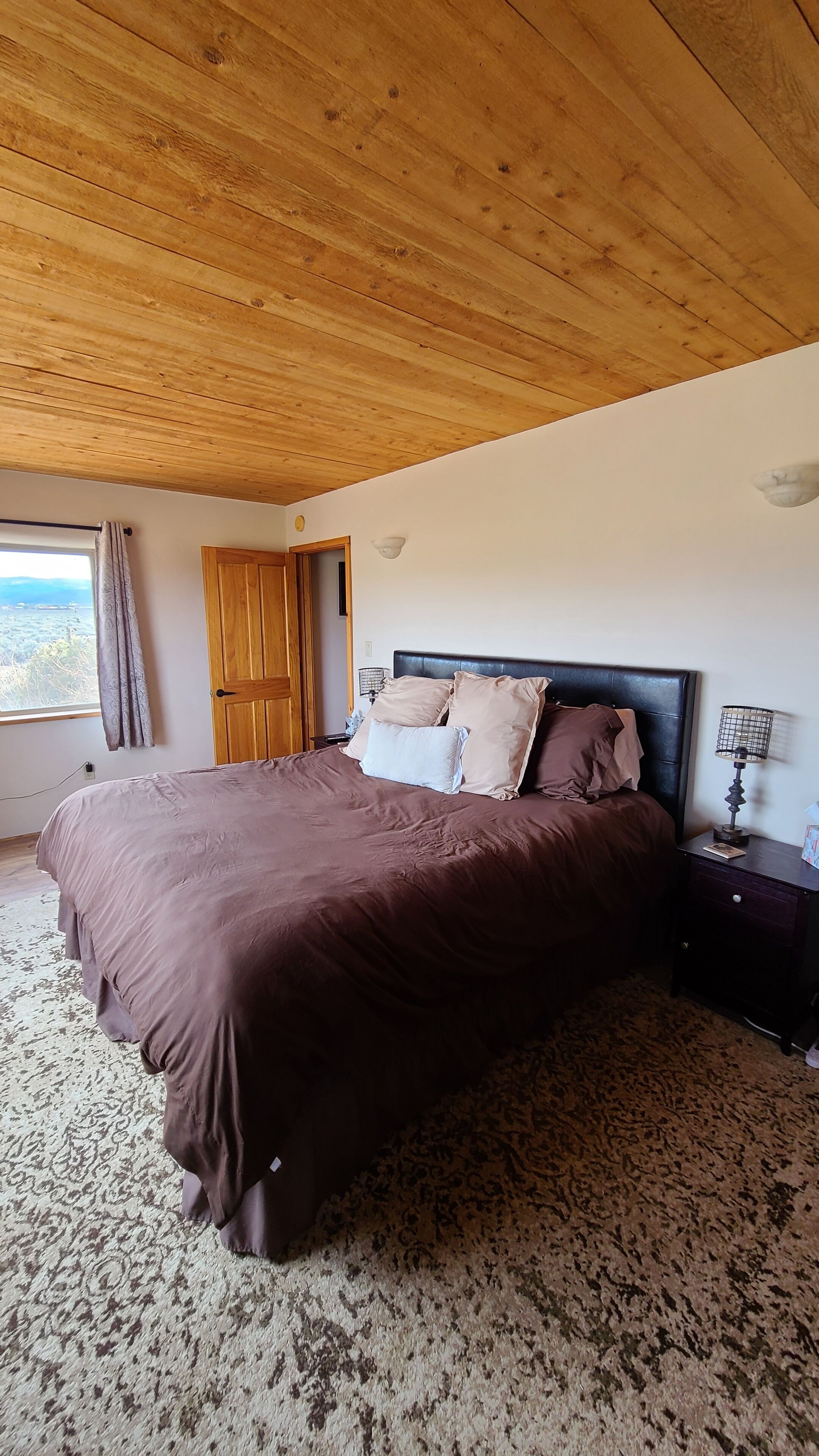 Un dormitorio con una cama grande, techo de madera, una mesita de noche y una ventana con cortinas.