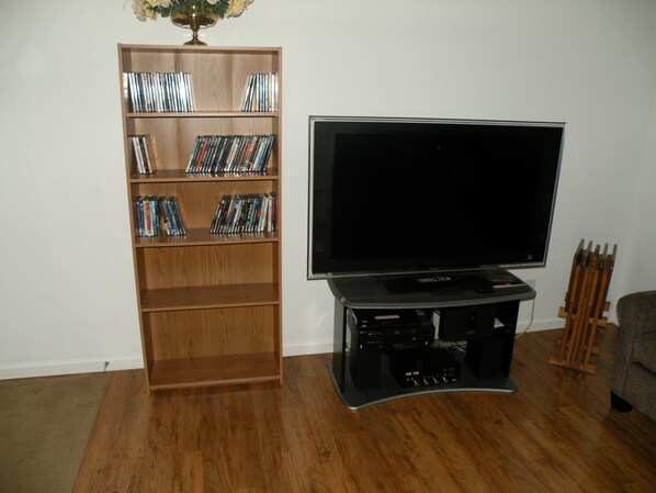 A living room with a wooden bookshelf filled with DVDs, a flat-screen TV on a stand, and a wooden chair.