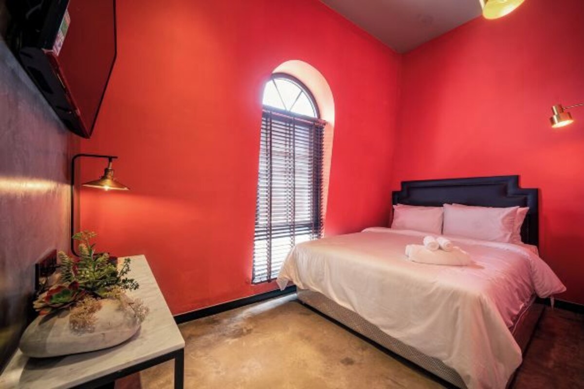 Une chambre avec un mur rouge, un lit avec une literie blanche, une petite table avec une plante et une fenêtre avec des stores.
