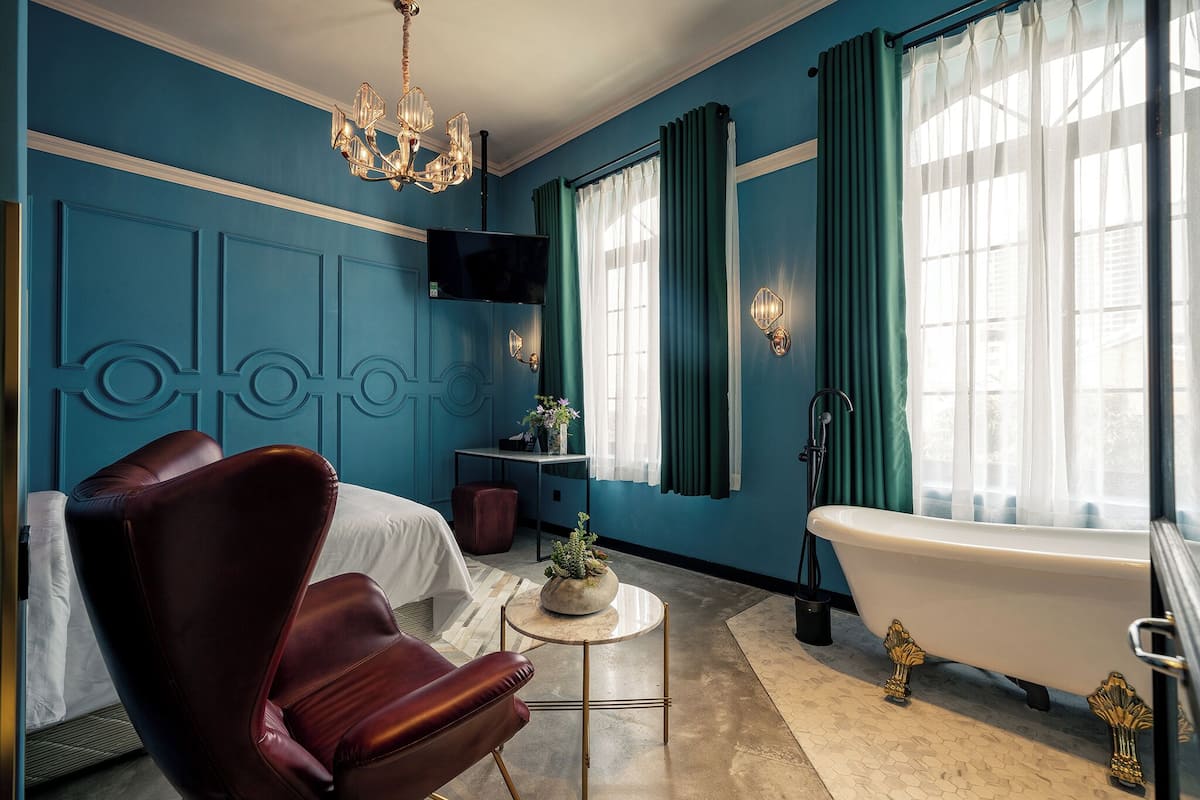 Une pièce comprenant un lit, un fauteuil en cuir rouge, une petite table ronde avec une plante, une baignoire blanche et une grande fenêtre avec des rideaux.