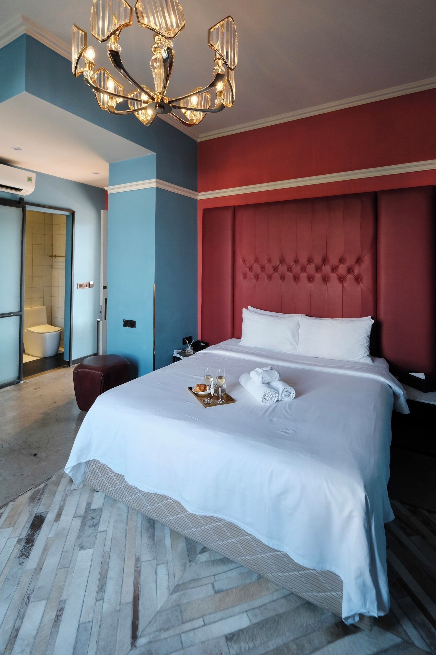 A hotel room with a large bed, a red headboard, a chandelier, and a bathroom visible through an open door.