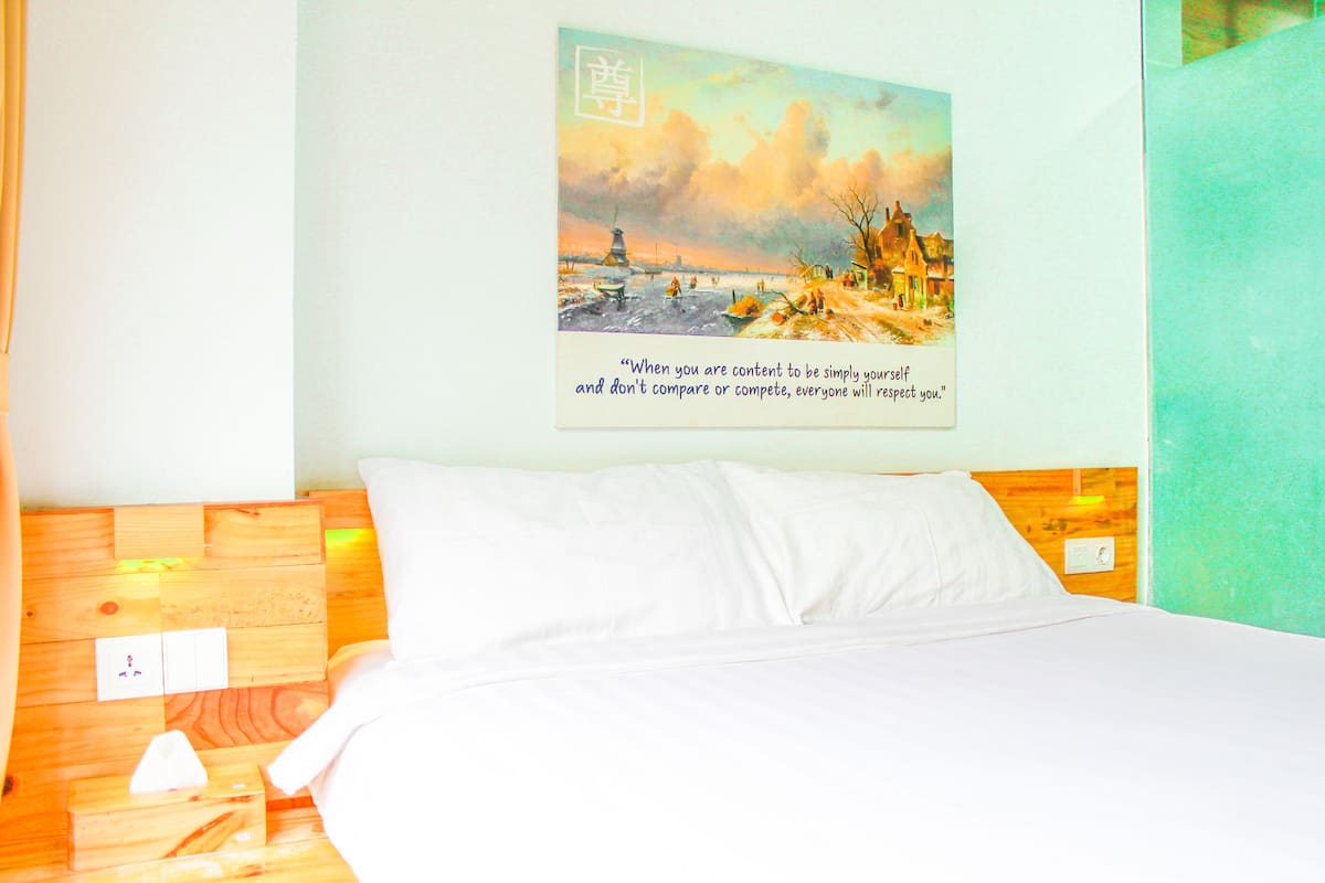 A hotel room with a wooden headboard, a bed with white linens, a wall-mounted painting of a coastal scene, and a small wooden nightstand with a tissue box and a remote control.