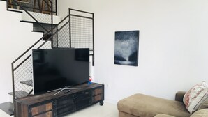 A living room with a large flat-screen TV on a wooden TV stand, a staircase with a metal railing, a beige sofa, and a floral pillow.