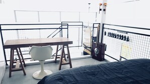 A room with a bed, a small table, a chair, and a view of the outside through a balcony railing.