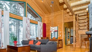Ein geräumiges Blockhaus-Interieur mit Holzboden, einer Sofagarnitur, einem Esstisch und großen Fenstern mit Blick auf den Wald.