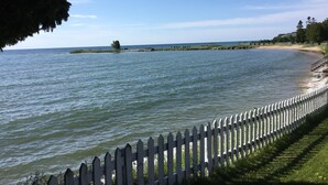 A coastal view with a white picket fence, calm water, and a distant shoreline with vegetation and a building. - Waterfront Home In Baileys Harbor ~ Fantastic Views Of The Harbor And Islands! (Baileys Harbor)