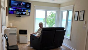A room with a bed, a massage chair, a TV displaying multiple channels, a small refrigerator, and a window with a view of the ocean. - Waterfront Home In Baileys Harbor ~ Fantastic Views Of The Harbor And Islands! (Baileys Harbor)