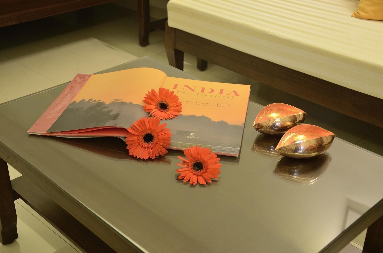 A book titled "INDIA the Beautiful" on a table with three orange flowers and two reflective objects.