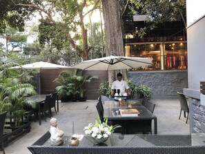 An outdoor dining area with tables, chairs, and umbrellas, surrounded by lush greenery and a building with a glass facade. - Hotel Ketan (Pune)