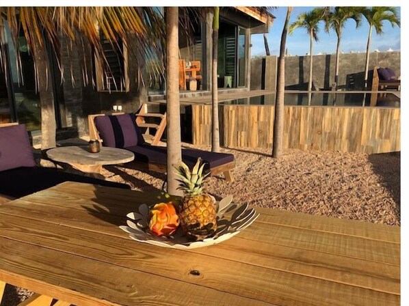 Een houten tafel met een ananas en een schaal fruit, omringd door terrasstoelen met kussens en een houten hek.