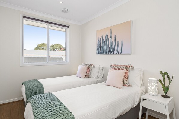 A bedroom with two beds, a window, a painting, and a nightstand with a cactus.