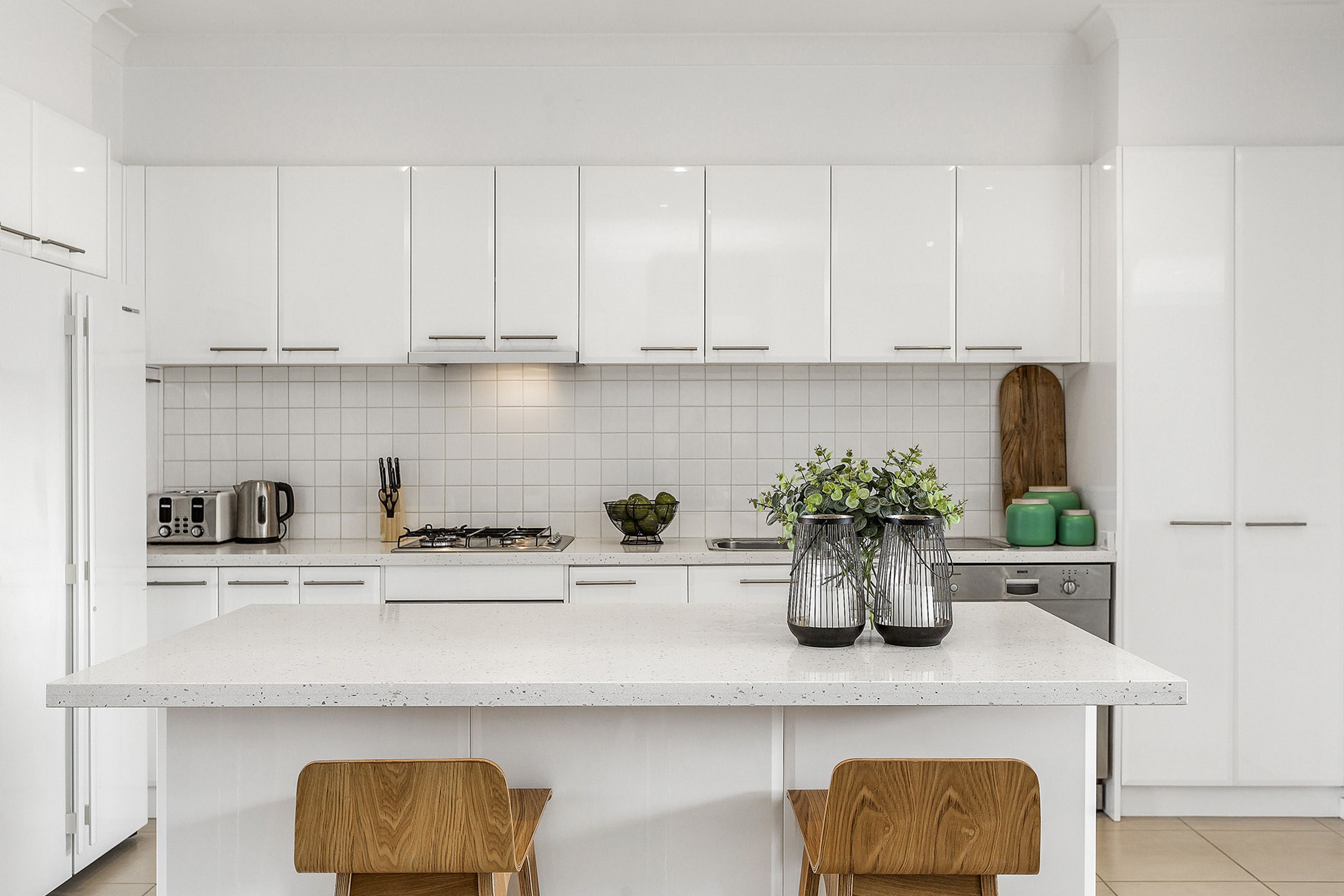 A modern kitchen with white cabinetry, a central island with two wooden chairs, and a Bosch dishwasher.