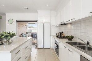 A modern kitchen with white cabinetry, a built-in oven, a sink, and a dining area visible through an open door. - The Richardson a  Dendy Apartment with Garage (Mentone)