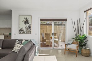 A living room with a grey sofa, a dining area with wicker chairs, and a sliding glass door leading to an outdoor patio. - The Richardson a  Dendy Apartment with Garage (Mentone)