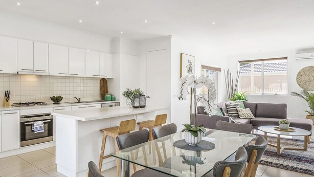 Une cuisine moderne avec des meubles blancs, un coin repas doté de tables et de chaises en verre, et un salon avec un canapé et des plantes décoratives.