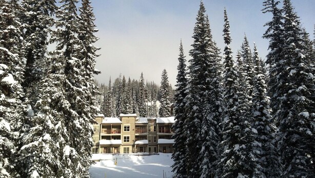 A snow-covered landscape with a multi-story building surrounded by tall evergreen trees. - Ski In/Ski Out Modern Condo in Wildwood  at Creekside (Vernon)