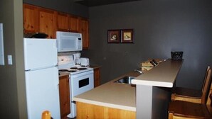A compact kitchen with wooden cabinets, a white refrigerator, a microwave, a stove, and a small dining area with a counter and two chairs. - Ski In/Ski Out Modern Condo in Wildwood  at Creekside (Vernon)