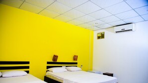 Ett hotellrum med två sängar, vita sängkläder, en gul accentvägg och en luftkonditioneringsanläggning.