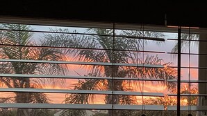 A window with horizontal blinds, through which a sunset is visible, featuring palm trees and a clear sky. - Coastal Studio Retreat!! WALK TO SURF & SAND or 1 MIN DRIVE!!! GREAT LOCATION!!! (Carlsbad)