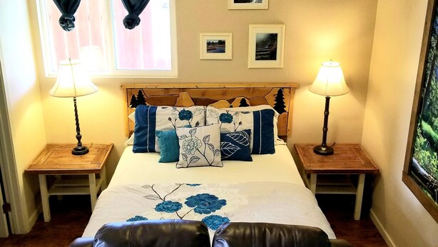 A hotel room with a bed, two bedside lamps, a headboard with a floral design, a window with curtains, and a small table with a lamp. - The Cottage on Campbell- In town studio with a view!  (Mariposa)