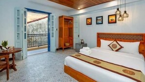 Une chambre d’hôtel avec un lit, une tête de lit en bois, une petite table avec une plante, une armoire et un balcon avec une balustrade.