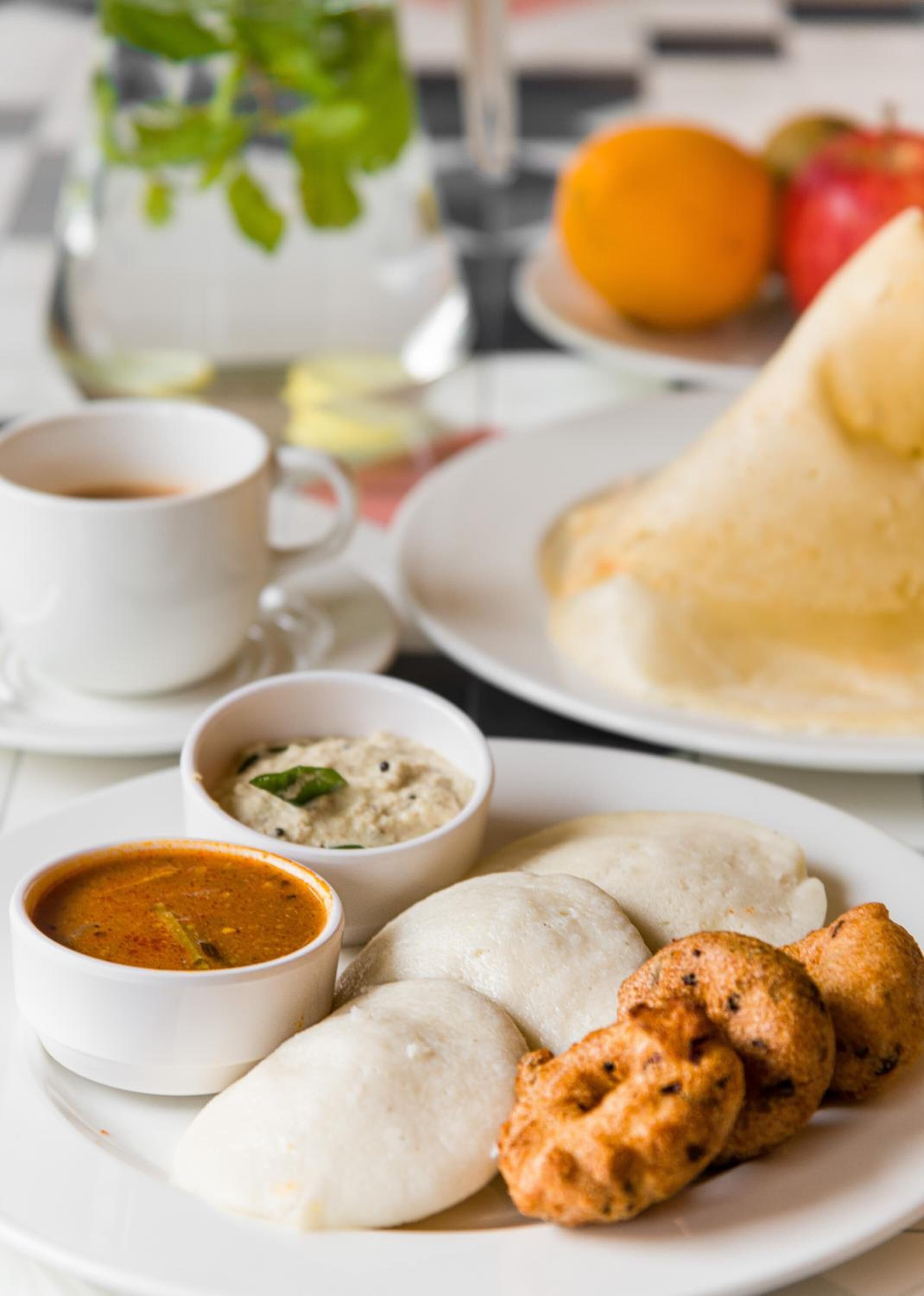Una mesa con una taza de té, un plato de idlis, un cuenco de sambar y un plato de vadas.