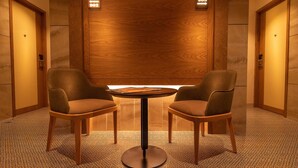 Een moderne lobby met twee leren stoelen rond een klein rond tafeltje, wanden met houten lambrisering en verlichte routeborden boven de deuren.