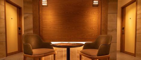 A modern lobby with two leather chairs around a small round table, wooden panel walls, and illuminated directional signs above the doors.