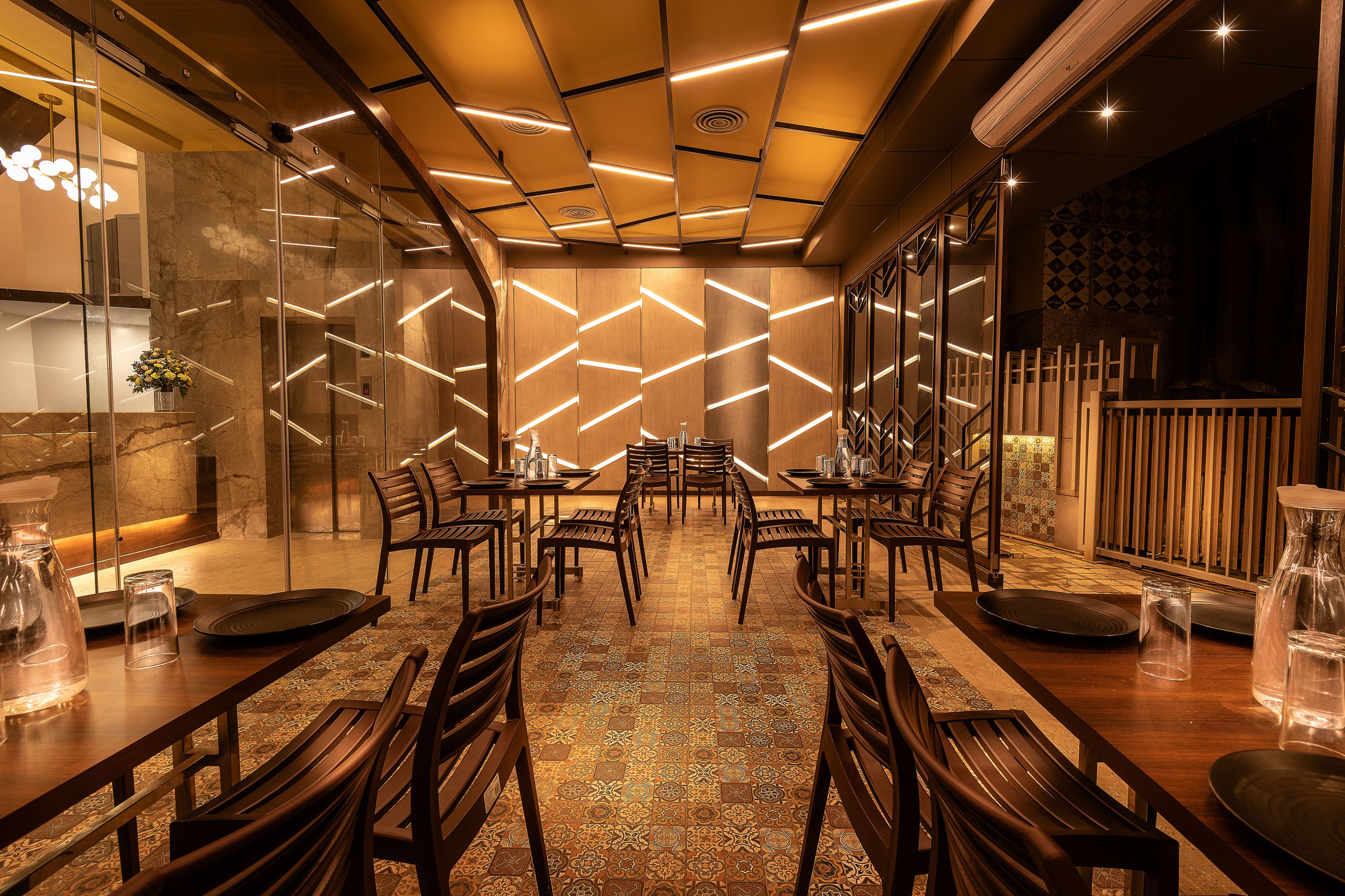 Un restaurante con un interior moderno, que presenta un suelo estampado, mesas y sillas de madera, y un techo decorativo con iluminación lineal.