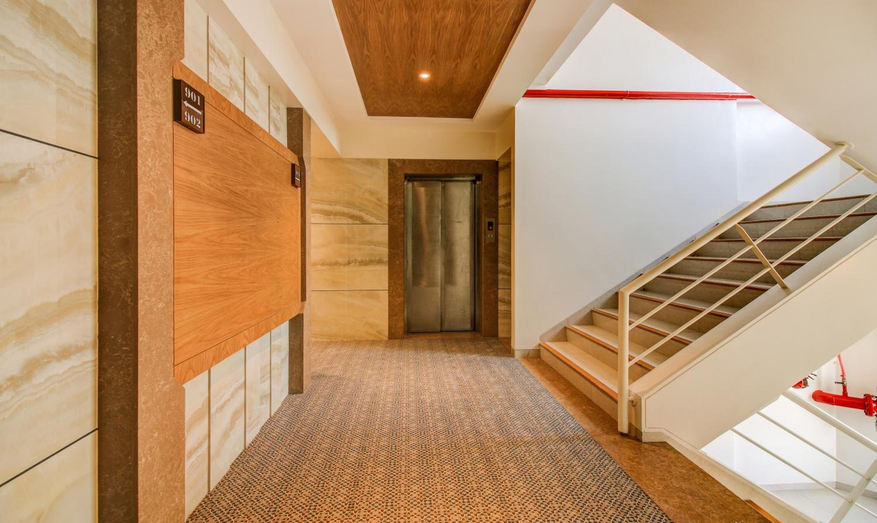 Passadís d'hotel amb paret de fusta, una catifes amb dibuixos, un ascensor i una escala amb barana de vidre.