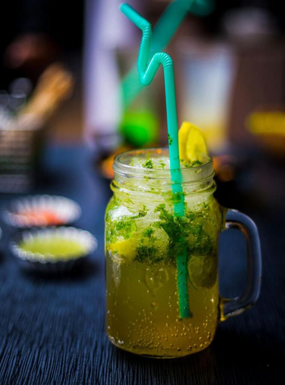 Un bocal en verre avec une paille verte, rempli d’une boisson verte et décoré d’une tranche de citron et de quelques herbes, posé sur une surface en bois sombre.