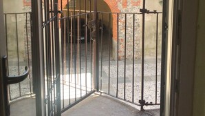 An open metal gate leading to a courtyard with a stone wall and a brick archway. - Secret Garden Art Museum (Arona)