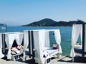 A row of four white canopy beds with a view of a calm sea and a distant hill. - Secret Garden Art Museum (Arona)