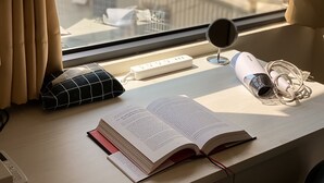 A desk with an open book, a hairdryer, a power strip, and a window with curtains. - Adagio Hostel (Taichung)