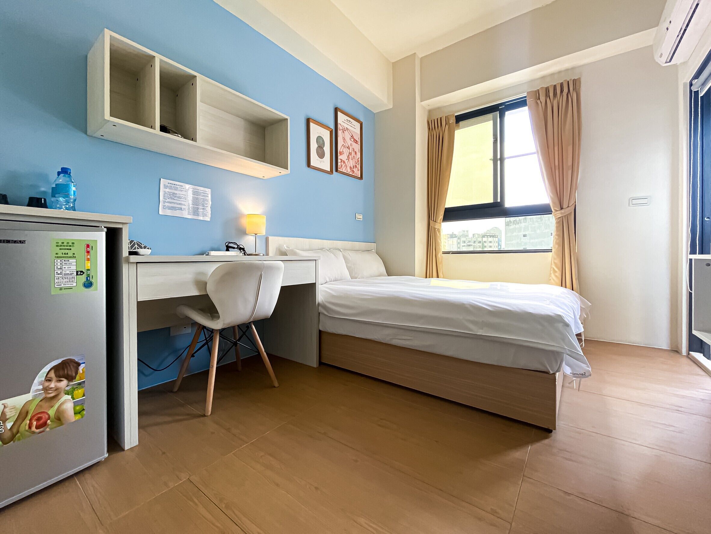 A compact hotel room with a bed, desk, chair, and a small refrigerator.