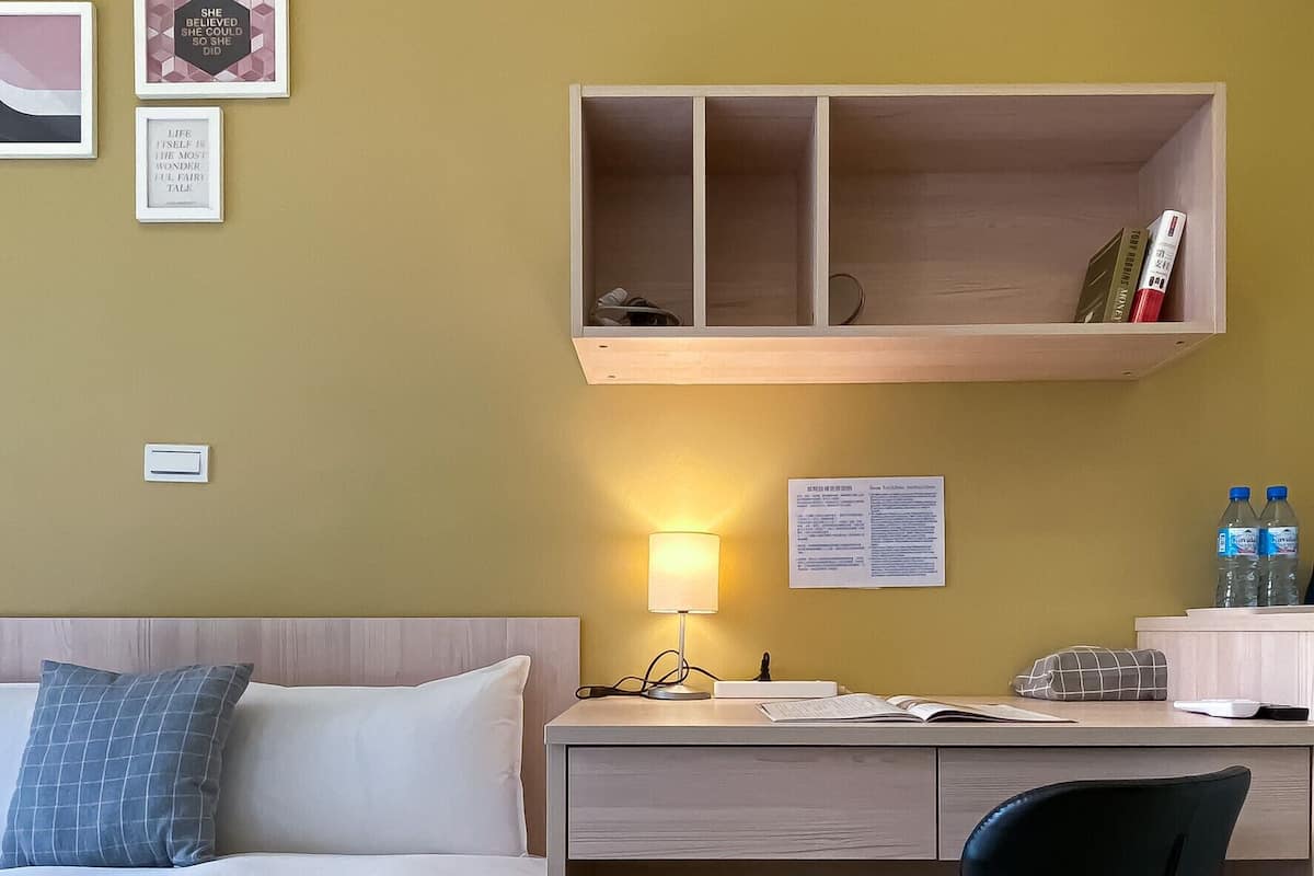 A hotel room with a bed, a desk with a chair, a lamp, and a shelf with books and a phone.
