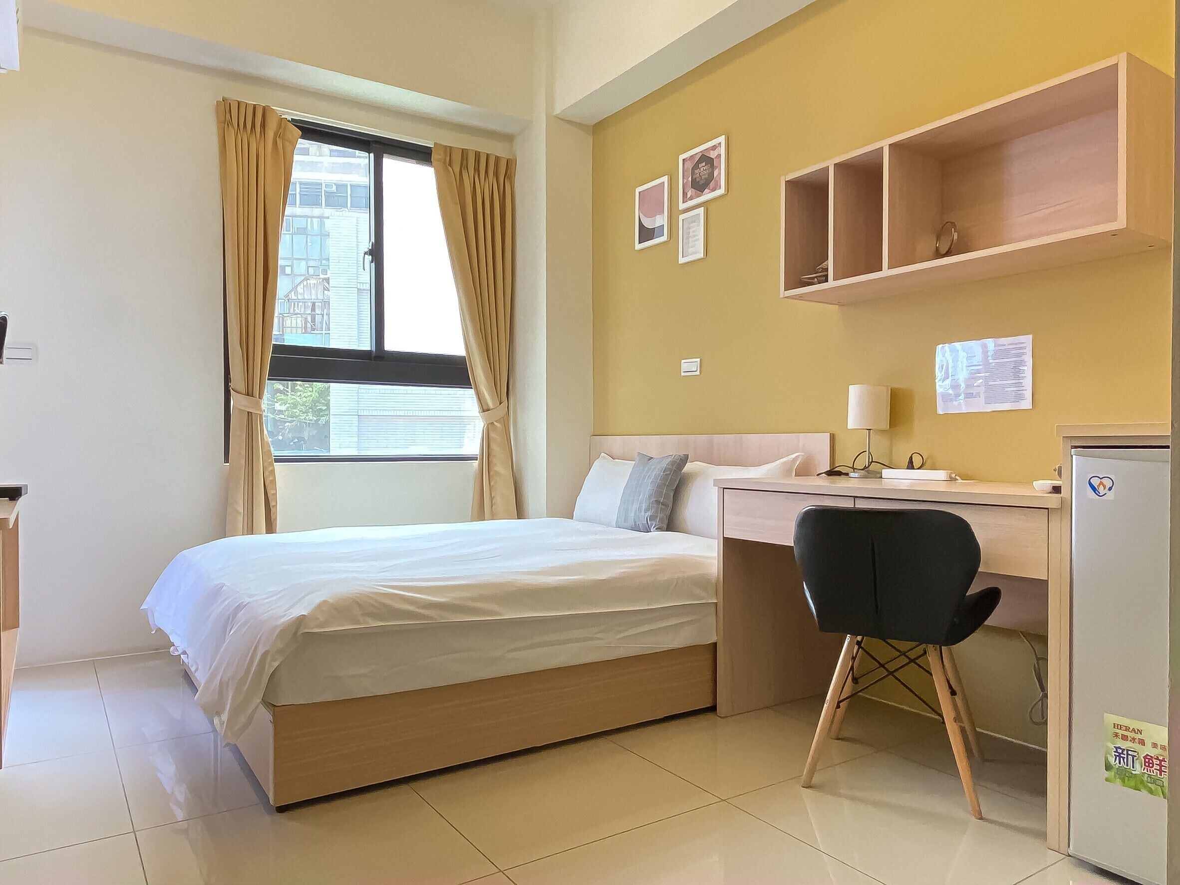 A hotel room with a bed, desk, chair, and a small refrigerator.