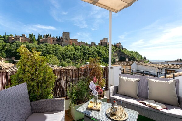 A balcony with wicker furniture, a vase with flowers, and a view of a historic city with castle-like buildings.