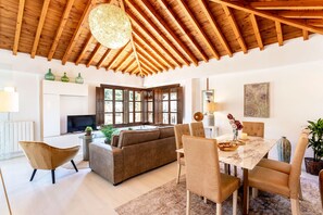 A modern living room with a wooden ceiling, a sofa, a dining table with chairs, and a TV.