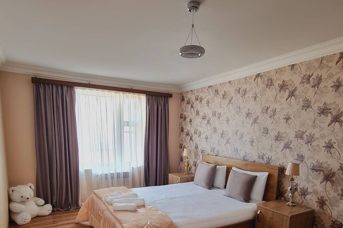 A bedroom with a large bed, wooden flooring, a teddy bear, and floral wallpaper.